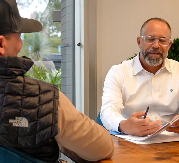 A man in a white shirt and glasses reviews and discusses paperwork with another person wearing a black vest and cap during a meeting at a wooden table near a window.