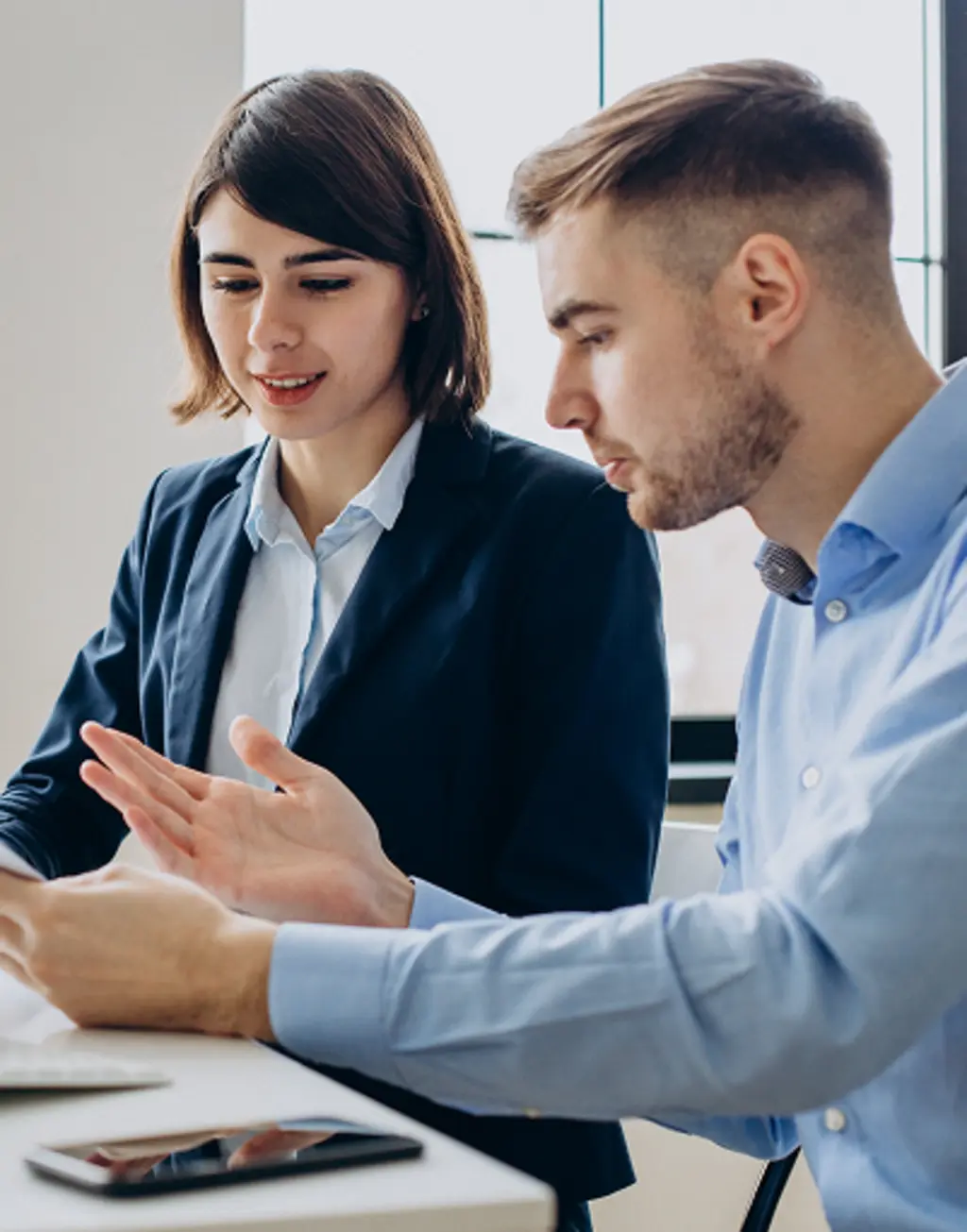 Two coworkers discussing a project at a desk in a bright office.