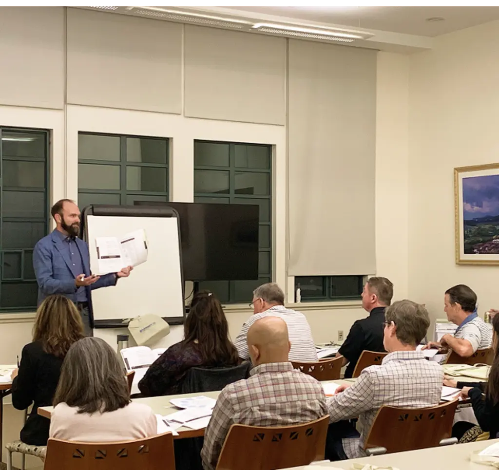 Instructor leading a classroom session for adult students while holding up a workbook at the front of the room.