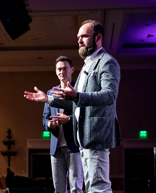 Two men in business attire speaking on stage at a conference, with one gesturing while addressing an audience.