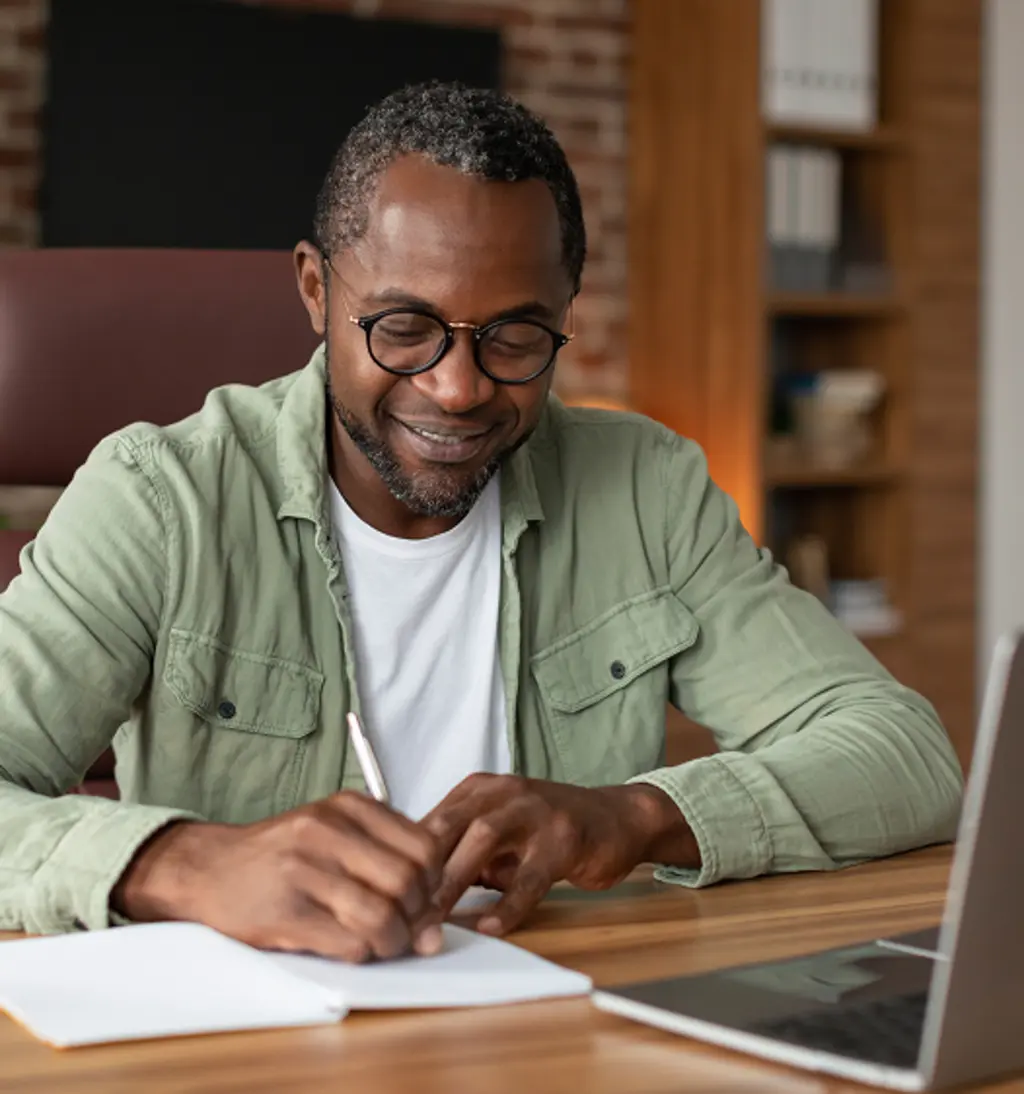 Man wearing glasses writing in a notebook while working at a laptop in a home office.