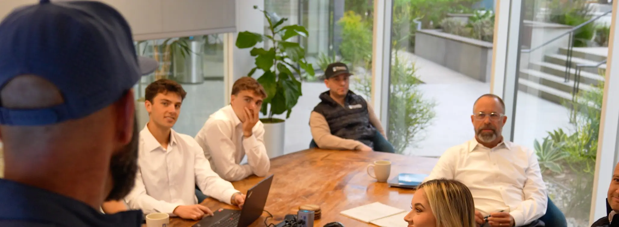 A group of four men sit around a wooden table in a modern office meeting room, listening to a person speaking off-camera; two wear white shirts, one wears a black vest and cap, and another has glasses and a beard, with plants and large windows in the background.
