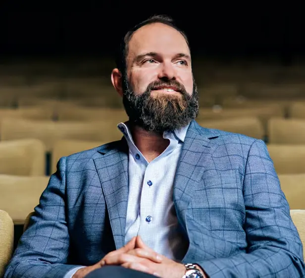 A bearded man in a blue checkered blazer and light shirt sits in an auditorium, smiling and looking off to the side with his hands clasped.