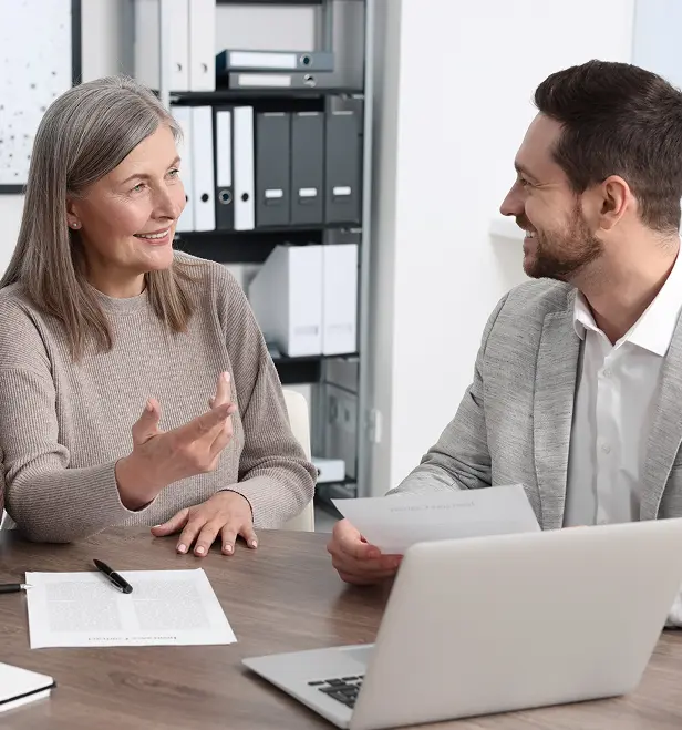 A middle-aged woman and a younger man sitting at a desk in an office, smiling and discussing documents with a laptop nearby.