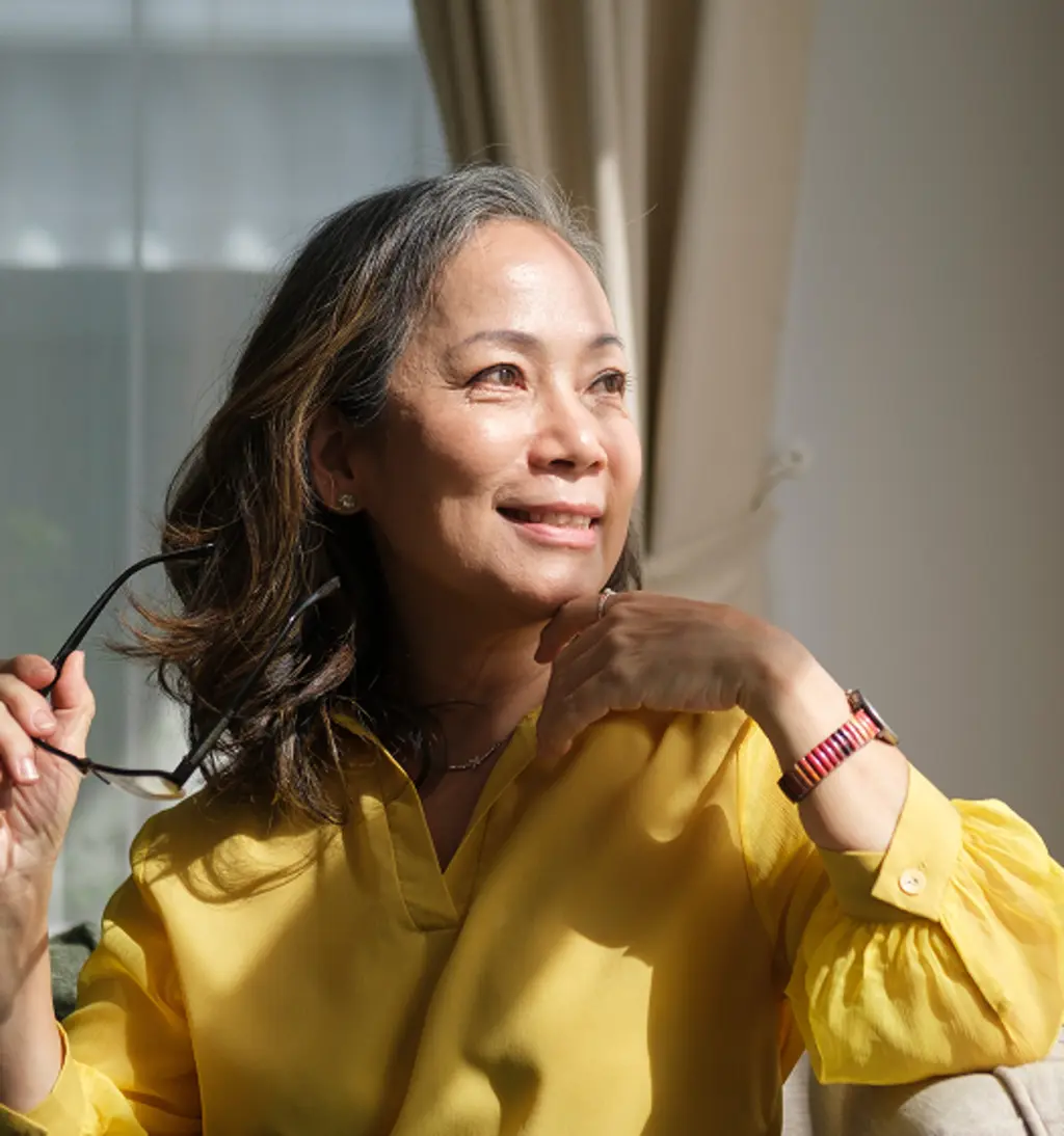 Woman in a yellow blouse holding glasses and smiling beside a sunlit window.