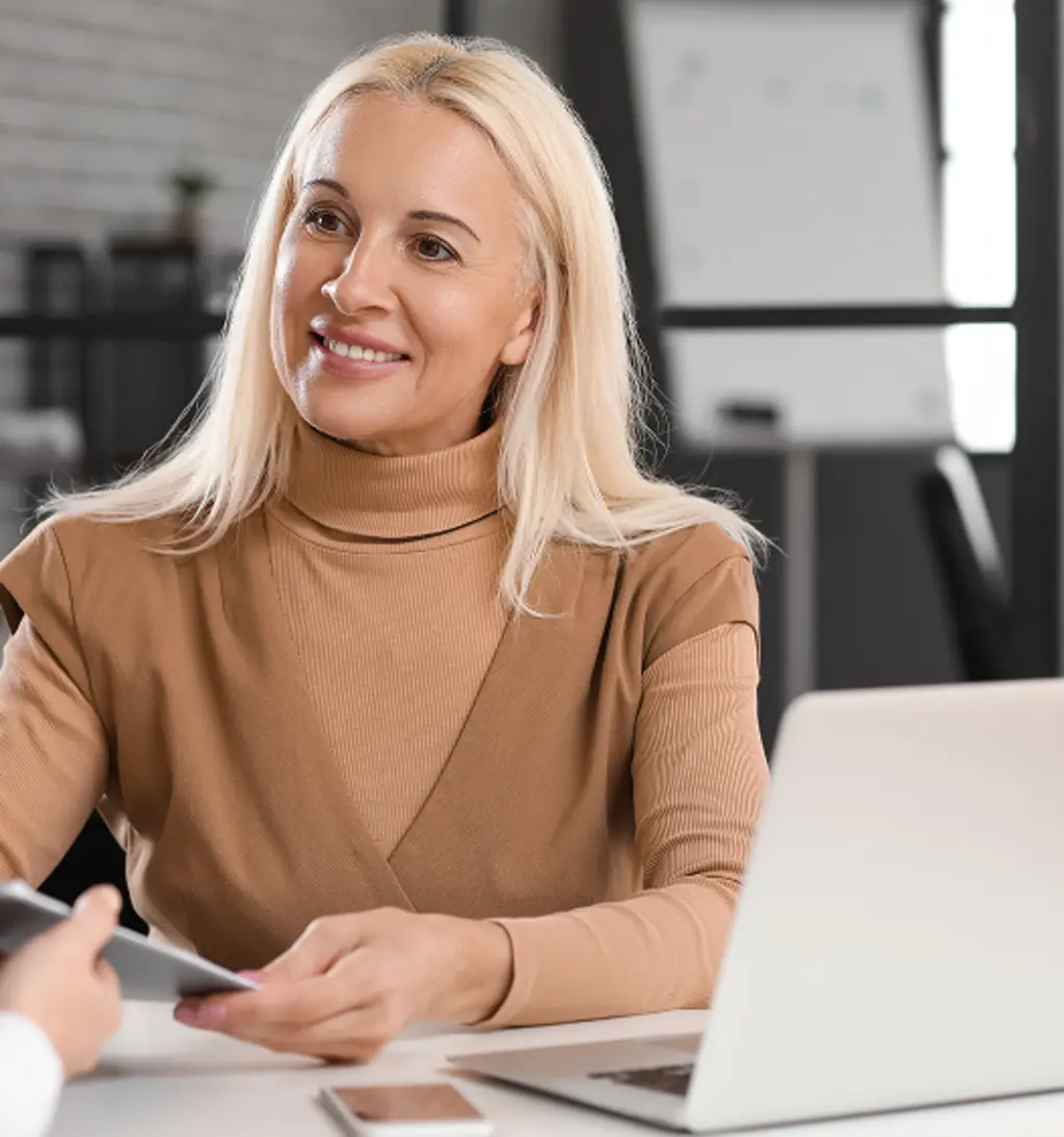 Smiling woman in a tan sweater meeting with a financial advisor at a desk with a laptop.
