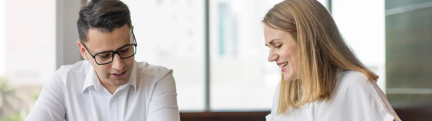 Two coworkers—a man wearing glasses and a woman—sit side by side in a bright office, smiling and looking down at documents as they collaborate.