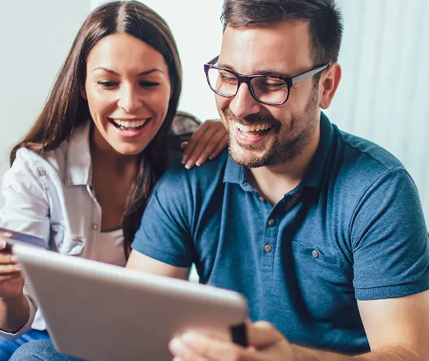 A smiling couple sitting close together at home, looking at a tablet while holding a credit card, appearing happy and engaged in online shopping or financial planning.