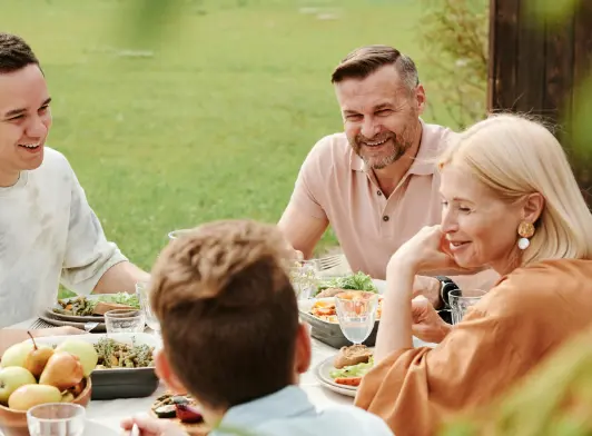 A group of adults and a child enjoying a meal together at an outdoor table in a garden, smiling and conversing in a relaxed setting.
