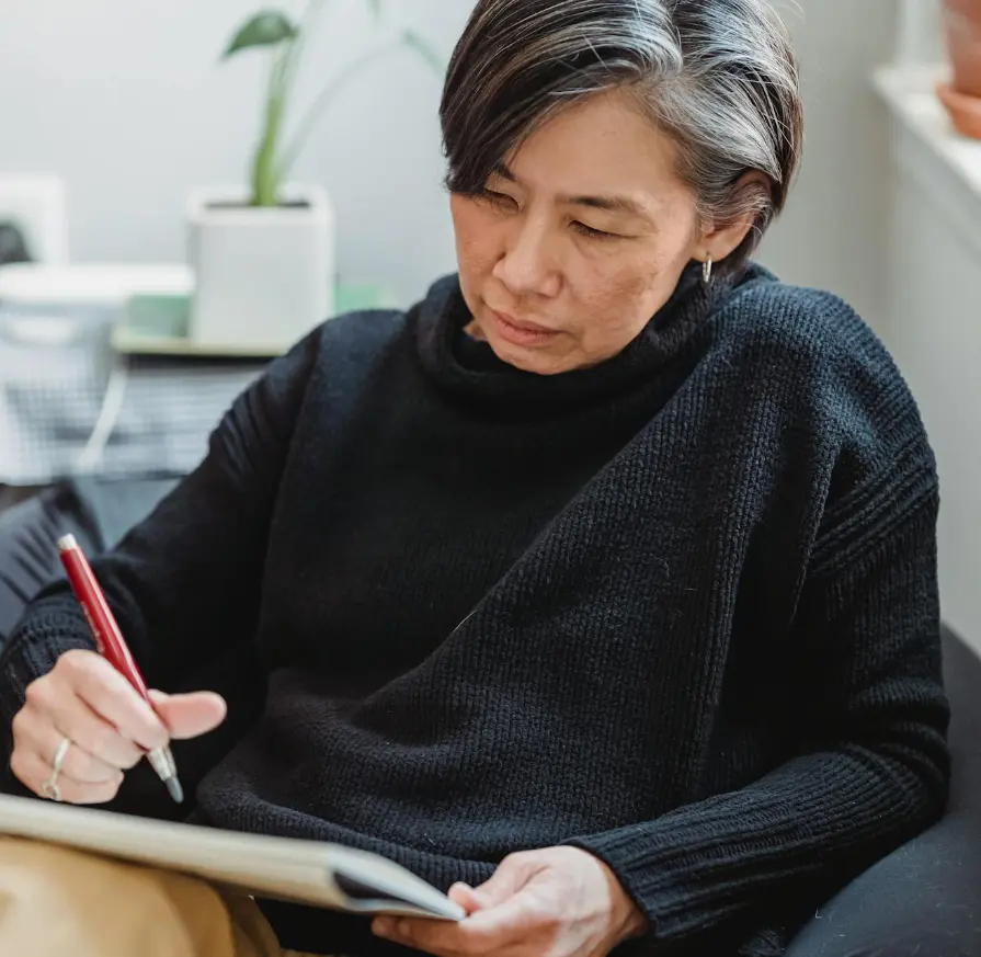 A woman with short gray hair sits indoors, focused as she writes in a notebook with a red pen.