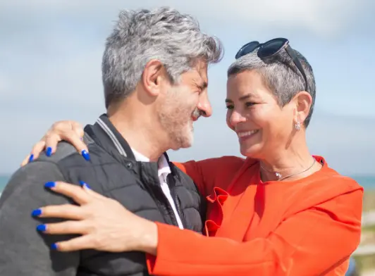 A middle-aged couple with short gray hair smiling warmly and embracing each other outdoors, with a soft sky and landscape in the background.