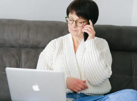 A woman wearing glasses sits on a couch using a laptop while talking on her phone, appearing focused and working from home.